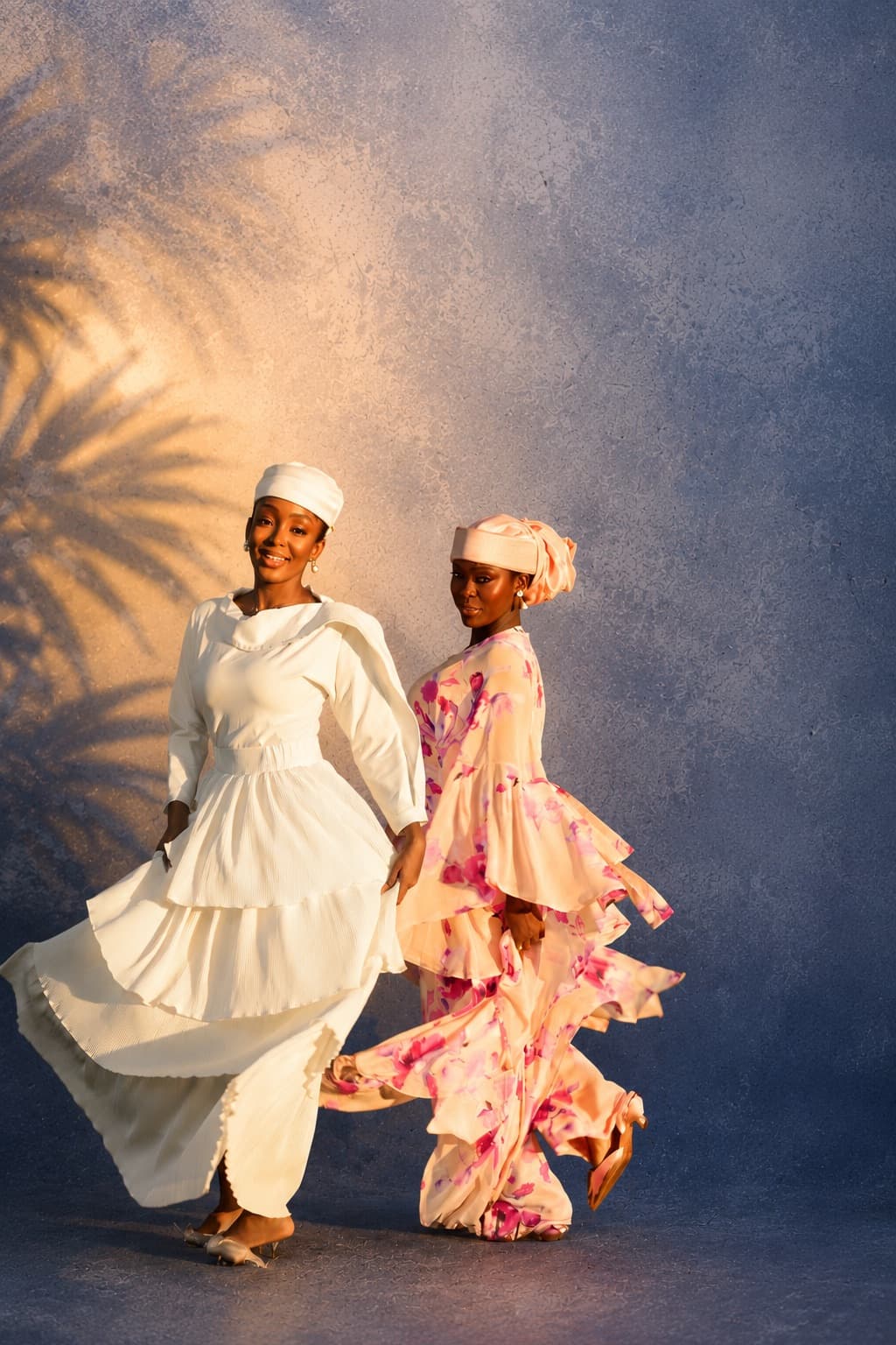 Two models in flowing ivory and blush floral dresses against a sunlit blue wall